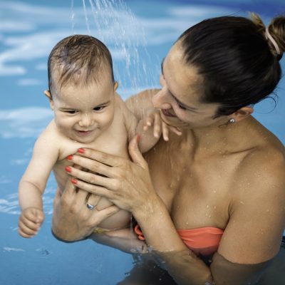 Instructor with baby boy and his mother playing in the pool. Spraying baby with watering can